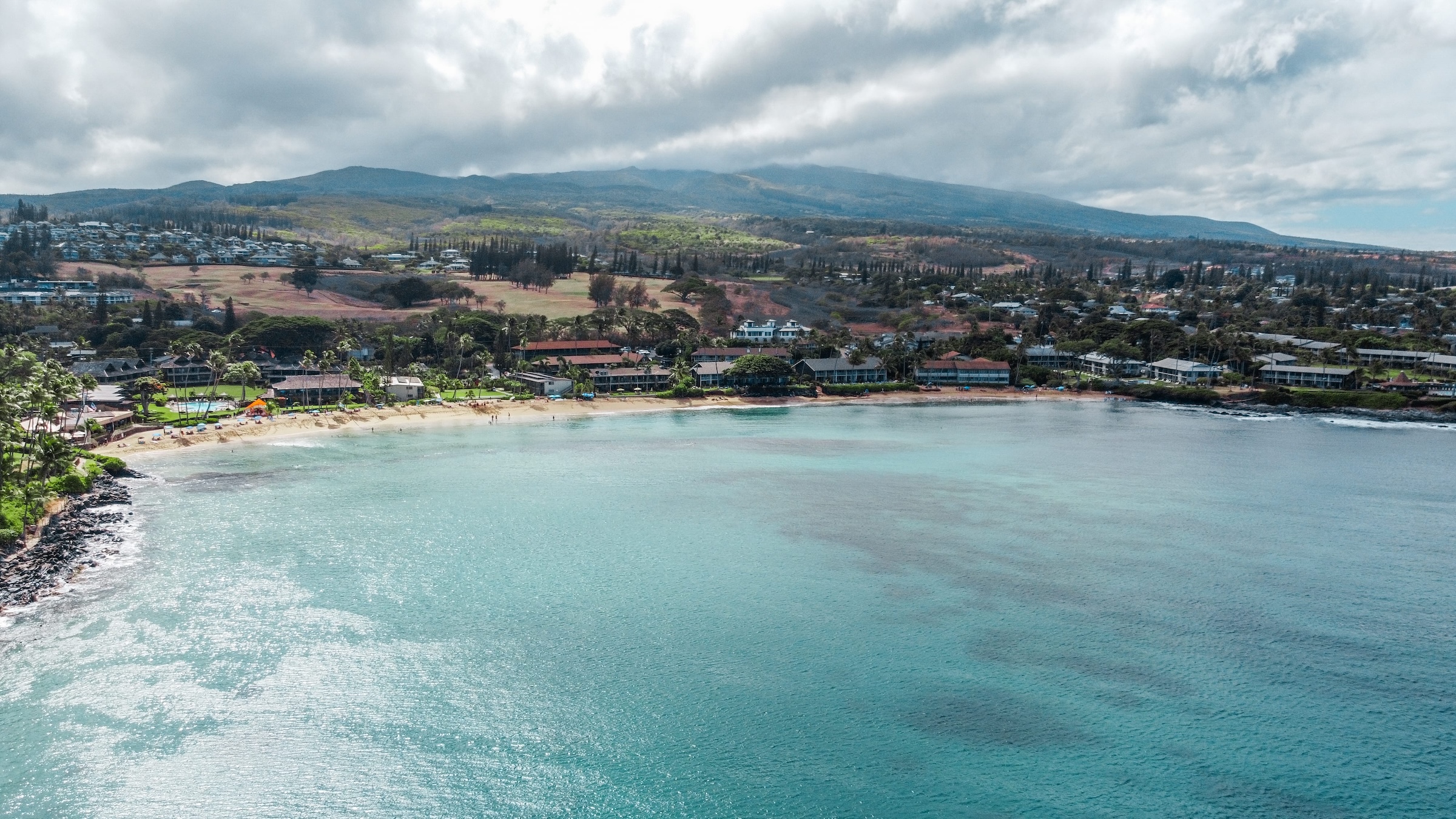 Napili Village Hotel, Aerial view of Napili Bay, Maui, showing the beach, clear water, and lush landscape, reflecting the 'Mālama Napili' restoration efforts.