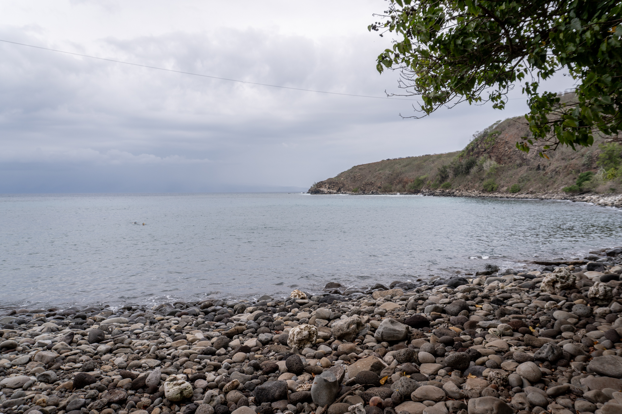 Napili Village Hotel, Rocky beach at Honolua Bay, Maui, with calm water and cloudy sky. Lush green hillside in the background.