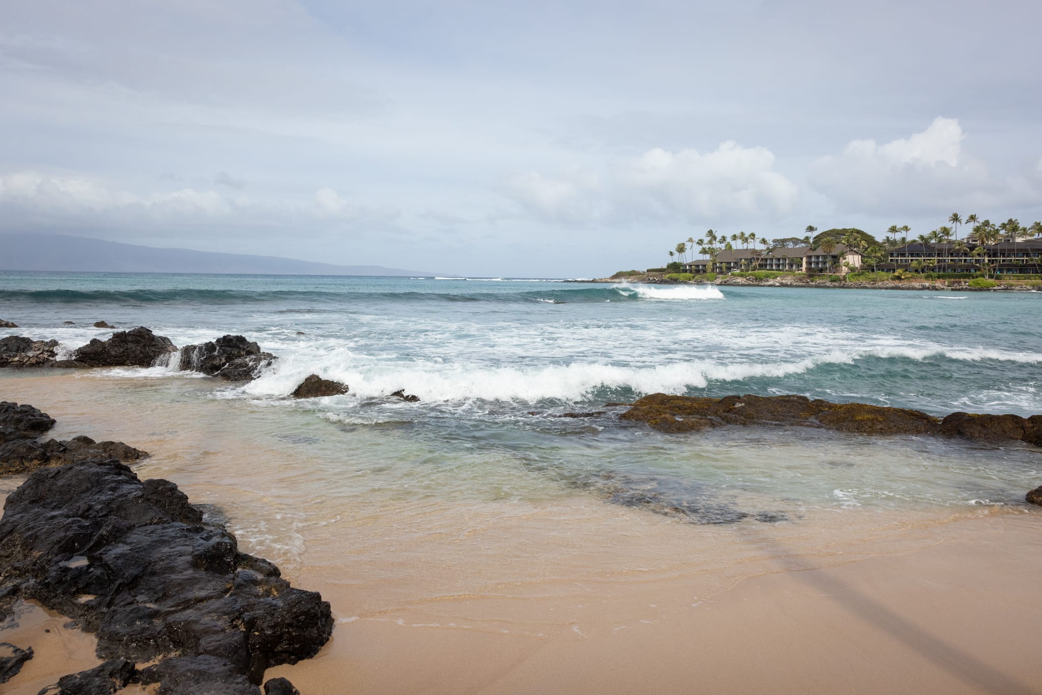 Napili Village Hotel, Waves breaking on the shore of West Maui, with buildings and palm trees in the background.